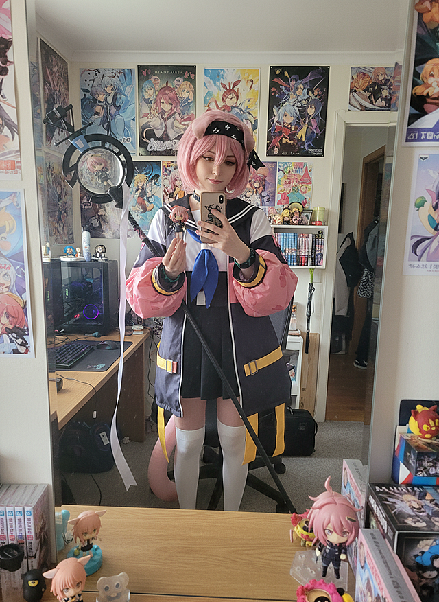 cosplayer真人照片，根据该图生成一张现实生活中扮演该角色的写实风格，人物所在的写实场景与原作相对应，人物一只手抬起拿着手机对着镜子正在自拍，另一只手靠近镜头托着带有与该cosplayer照片相对应的玩偶手办。