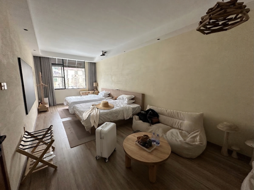 把深灰色行李箱更换为白色带轮子行李箱，在靠近镜头的床头放一块浅色格纹丝巾，在丝巾旁边放一顶草编宽檐草帽，保持整体原木简约风格协调统一预览效果