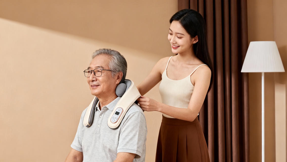 温馨居家场景的电商产品宣传图，暖棕米色为主色调，柔和自然光营造治愈氛围。画面主体是一位面带温柔微笑的年轻东亚女性，身穿米色吊带衫与棕色半裙，正贴心地为戴眼镜的年长父亲（身穿浅灰色短袖衬衫）佩戴浅米色颈预览效果