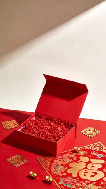 一张具有高度真实感的产品摄影风格图像，主体为一个打开的方形红色礼盒，内部填充着大量鲜艳的红色碎纸丝带，呈现出蓬松饱满的质感。礼盒放置在红色的平面上，周围散落着金色边框的“福”字剪纸，其中一张较大的剪纸展开着，上面有传统的龙纹和花卉图案。画面中点缀着几个小巧的金色元宝，增强节日氛围。整体色调以正红色和金色为主，色彩对比鲜明强烈，喜庆感十足。光线从斜上方照射下来，在礼盒和剪纸表面形成明亮的高光和清……预览效果