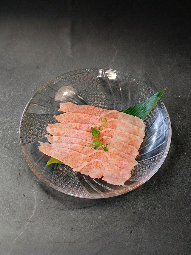 超高清专业美食摄影，8K画质，极致细节。参考花蛤菜品母版的摄影风格，将这道雪花肥牛刺身/涮涮锅牛肉菜品进行统一修图。要求：1）严格保留原图中的所有菜品内容：水晶玻璃盘、七片雪花牛肉、竹叶、豆苗装饰，食材和摆盘完全不变，保留玻璃盘的菱形刻花纹理；2）背景替换为深灰黑色哑光石材/水泥质感桌面，带非常细微自然不规则的纹理，参考花蛤母版的背景亮度和纹理强度，真实桌面材质感；3）统一为柔和侧上方主光，食物主体清晰明亮，背景压暗，牛肉脂肪纹理通透有光泽，玻璃盘有自然通透反光和边缘高光，底部产生真实接触阴影，阴影方向与花蛤母版一致；4）统一45度俯拍视角，竖版构图，玻璃盘位于画面偏下区域略偏左，上方保留大面积深色负空间，左右及底部留白比例参考花蛤母版；5）统一色温、对比度、黑位、饱和度，整体餐饮摄影风格与花蛤母版高度一致，肉类呈现自然暖红色调；6）极致高清：牛肉的大理石脂肪纹理、肉的细腻肌理、玻璃盘的刻花纹理和通透反光、竹叶的叶脉全部清晰可见，专业商业美食摄影级画质；7）禁止改变菜品种类、禁止添加花蛤或贝壳、禁止纯黑背景、禁止塑料质感、禁止插画感、禁止文字水印。