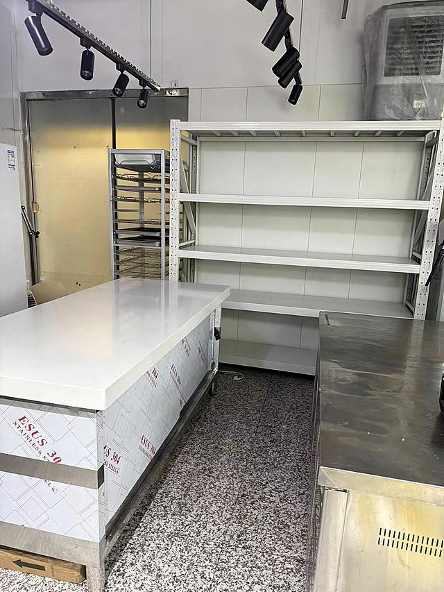 去除后方白色多层货架上的所有物品（包括纸箱、塑料盒、包装袋、瓶罐、篮子等所有摆放在货架各层上的物品），仅保留空的白色货架主体结构，货架的金属框架和层板完整保留。严格保持房间内其他所有主体、细节、结构、颜色、光线、透视角度完全不变，不得添加或修改任何其他元素。去除物品后的区域用周围真实的墙面和货架结构自然填充，光影和材质与原图一致，过渡自然无痕。最后整体提升图片清晰度，画质自然真实，保持原照片的真实感。