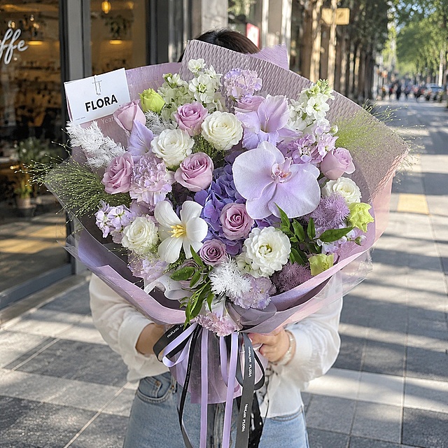 以图中的花束为主体，严格保留花束中所有原有花材（紫玫瑰、淡紫蝴蝶兰、紫色绣球、白色水仙百合、白色小花、紫色小花、绿色花材、紫色包装纸、FLORA卡片、丝带等）的种类、数量、颜色、形态、位置和包装完全不变，仅在花束间隙中间插添加5朵白色洋桔梗（白色杯状花瓣），自然点缀分布。去除原图中的人物和室内黑色背景。画面为白天城市街拍场景，一位中国年轻女性正面抱着这束花，不露脸（头部被花束遮挡或裁切在画面外），自然站立姿态，身穿简约日常服装。背景为白天明亮的城市街道街景，有咖啡店橱窗、人行道、柔和阳光，背景适度虚化突出花束。整体风格清新自然，写实摄影质感，花束为视觉焦点。严格锁定宽高比为1:1。