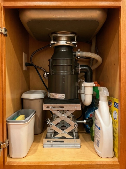 俯拍视角，在一个小巧但功能齐全的公寓厨房中，水槽下方空间狭小。垃圾处理器升降台以最紧凑的结构安装其中，巧妙地将处理器托举并收纳。镜头展现其如何最大化利用有限空间，画面温馨、高效。比例3:4预览效果