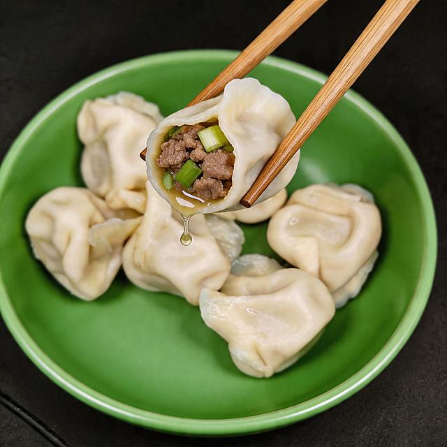 添加筷子夹起一个饺子，夹起的饺子露出牛肉和鲜亮的大葱内馅，有汤汁流出，整体画面垂涎欲滴很有食欲，保留绿色碗和其他饺子不变