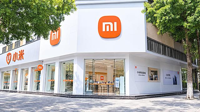 精修这张小米门店照片，去除画面中所有杂物：移除左侧的人物、电动车及紫色遮阳棚，移除右侧的三轮车、电动车、交通标志牌、路牌，移除空中的电线，移除地面的石球；保留门店建筑主体和两侧的树木，提升画面整体亮度，让光线明亮通透，色彩干净清新，门店外立面洁白如新，玻璃门通透干净，地面整洁，整体呈现出崭新开业的干净明亮效果，画质高清细腻，真实摄影风格