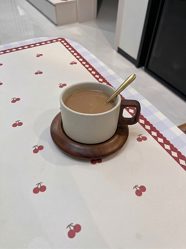 将原图中的黑色杯子和木质杯垫替换为参考图中的米白色粗陶杯子搭配木质杯垫，保持杯子和杯垫的角度与原背景桌面协调，主体不发生严重形变变高拉长，还原杯子和杯垫的原始样式，保留桌子上的樱桃图案桌布和金色勺子