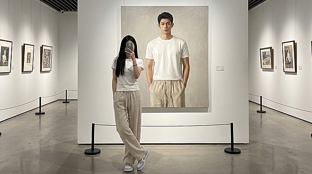 一位清冷气质的18岁东亚年轻女生她留着黑长直发型，身穿纯白色短袖上衣，下身搭配浅米色亚麻休闲裤，脚踩浅灰色耐克板鞋，她身后是一幅等身大正方形的男性写实油画，画中人物穿着同款上衣，下身搭配同款亚麻休闲裤，双手插兜，与女生形成呼应式穿搭。展厅两侧墙面挂有小幅艺术画作，地面设有黑色隔离立柱，女生用左手持手机挡脸自拍，保持不露脸，微微侧身，双腿轻交叉，整体氛围清冷高级，兼具艺术感与穿搭展示性，光线柔和且层次分明，突出人物与画作的互动感
