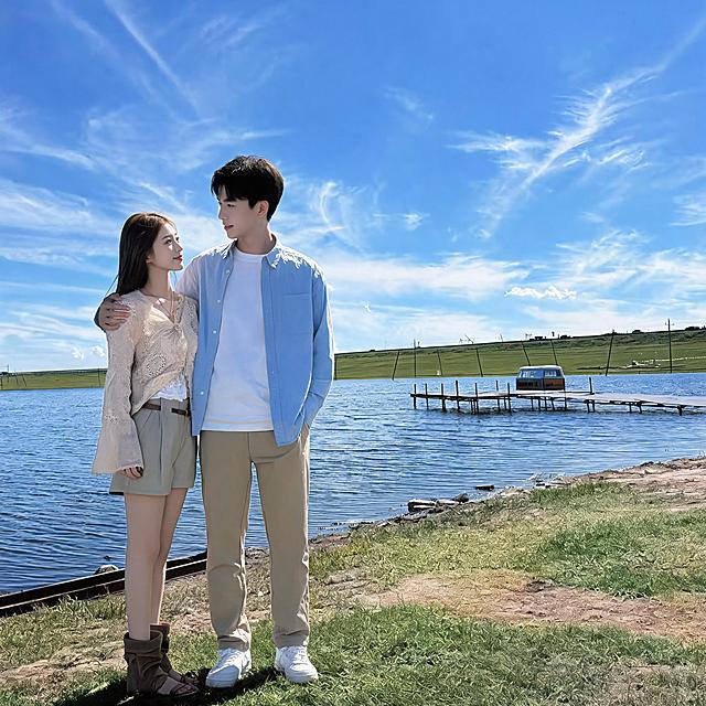 生成日系胶片清新风情侣双人合影写真（单幅画面，无拼图），保留参考图中女生的脸型、发型、年龄、五官特征，相似度99%，新增游泳运动员张展硕形象的年轻男生（帅气阳光、短发、身材健硕），两人五官高度还原，忽略参考图中的墨镜配饰。低饱和清透蓝调，富士胶片质感，色彩干净通透，柔和自然光，发丝逆光轮廓光，面部无硬阴影，大光圈浅景深，虚实交织的朦胧感，干净治愈松弛自然；女生清透奶油肌，真实皮肤纹理，白皙通透，细腻无瑕，棕色长直发自然垂落，穿米白色镂空针织开衫内搭白色吊带，卡其色短裤，咖色堆堆靴；男生清爽干净肤质，自然肤色，利落黑色短发，穿浅蓝色休闲衬衫搭配白色T恤，卡其色休闲长裤，白色运动鞋；两人并肩站在湖边草地上，男生单手轻轻搭在女生肩膀上，女生侧身依偎在男生身旁，两人相视微笑，眼神温柔充满爱意，甜蜜恩爱互动；背景是开阔的蓝色湖泊，远处绿色草坡与白色风力发电机，蓝天白云，水面波光粼粼，木质栈桥延伸至湖中，岸边青草随风摇曳；中景构图，三分法构图，人物居中偏左，背景层次丰富；分辨率4k，电影级光影质感，商业人像摄影级别，画面比例1:1