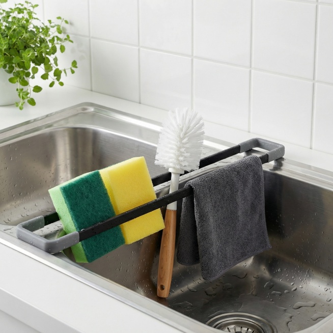 严格保留图1厨房水槽收纳架，外观、尺寸，放置在不锈钢水槽上，海绵刷放置在架子上、悬挂清洁布、木质手柄的白色清洁刷头卡在架子中间，背景是白色瓷砖墙面，画面右侧，左上角有一株绿叶植物点缀。整体光线明亮，营预览效果