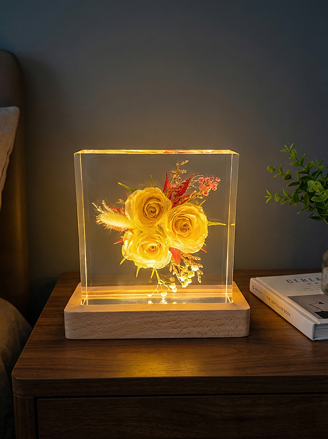 保持产品主体结构不变，不改变黄玫瑰干花与原木底座的造型，保留透明方形树脂结构，树脂部分清亮透明犹如玻璃，画面干净无浮尘，产品只出现一次。放置在深色胡桃木床头柜表面，深灰蓝色卧室墙面背景，夜晚氛围，LED暖光从原木底座透出，整体温馨氛围感，暖黄色调，柔和漫射暖光，远处背景虚化。商业摄影，温暖底部柔光，突出透明树脂内部花朵。居中构图，平视拍摄。旁边放一本合上的软装书，点缀一小枝绿植。商业级4K超清画质，画面通透氛围感强。保持3:4画面比例。西方面孔,金发,深眼窝,雀斑,杂乱房间,死黑阴影,硬光,乡村风,高对比度,欧美妆容,变形,扭曲,悬空,比例失调