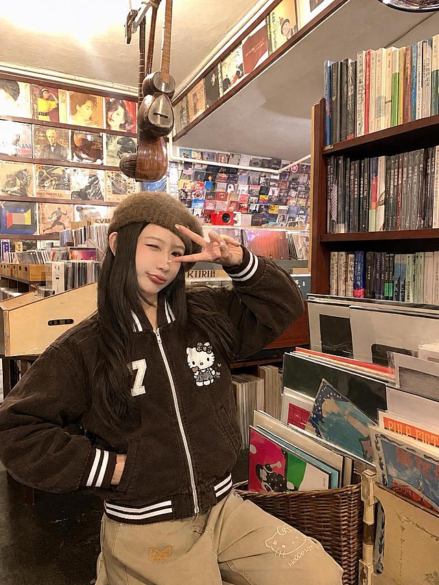 将图5的深棕色Hello Kitty棒球夹克穿到图4模特身上，严格复刻衣服版型、花色、纹理、面料、数字7刺绣、Hello Kitty图案、白色条纹罗纹领口袖口下摆、拉链设计，衣服贴合身体，服装褶皱自然，真实人像摄影，自然光，皮肤质感真实，高清，写实，柔和环境光影，无水印，无文字，无logo，无畸形肢体，画面干净，不要改变人物长相，保留原图背景和人物姿势