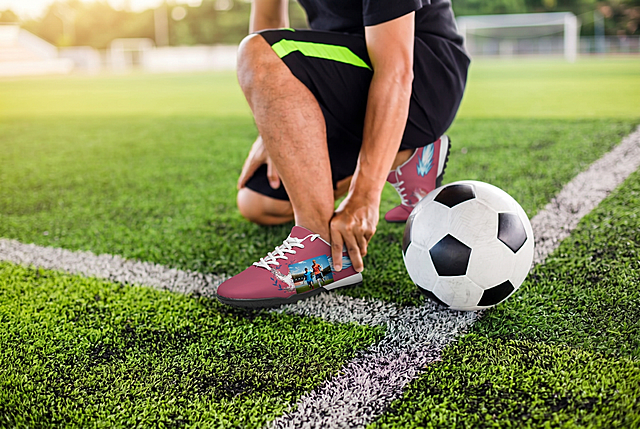 参考图1的风格和构图，将图2这款玫红色定制足球鞋放在足球场草地上，人物单膝跪地手扶脚踝，旁边放一个足球，保留图2足球鞋原有的玫红色底色、飞翼图案和双人合影印花，真实户外摄影，暖调阳光质感，和图1风格一致