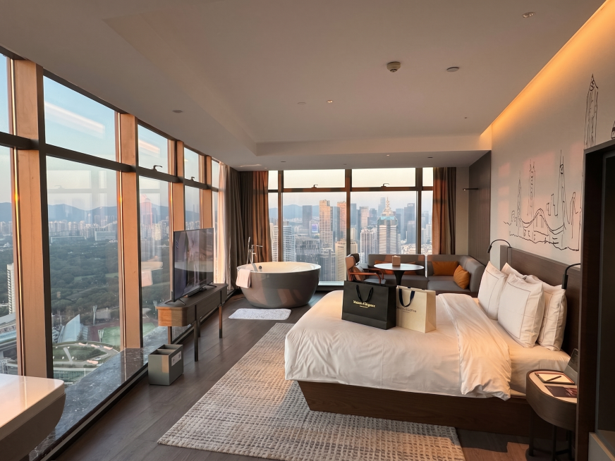 在当前城市景观豪华卧室图片中：1）在床前区域铺设一块地毯；2）在床上放置两个购物袋，自然摆放；保持原浴缸、沙发、电视及城市全景视野不变，风格现代高级预览效果
