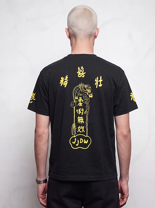 请将参考图1的【黑色短袖T恤】1:1还原其款式、颜色、材质、剪裁、图案、版型、印花细节、logo等所有细节，穿到参考图2的模特身上；完全保持参考图2模特的身姿、发型、气质、动作/姿态不变（含手部位置/朝向/重心/肩线）；模特身上参考图1未覆盖的部位（裤子、配饰、手表等）严格保持参考图2原样；画面背景/光影/构图/景别严格保持参考图2原样。

服装以参考图1为准，完整保留款式、颜色、材质、剪裁、图案、版型、印花、logo等一切细节。参考图1未覆盖的所有部位——【裤子、配饰、手表、内搭等】——严格保持参考图2（模特图）原样，保持原有画面内容与细节一致。

超写实商业摄影质感，高级时装大片风格，画面干净高级，细节丰富真实，色彩还原准确，整体质感细腻。