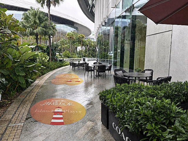 在现有基础上再多添加两组同款黑色编织户外桌椅（配灰色坐垫和深灰色圆桌），沿步道向远处方向依次摆放，整体布局自然合理，透视角度与空间比例准确。同时优化地面的两个地贴：调整地贴的透视使其完全贴合地面弧度，增加地贴的真实质感，呈现地面贴纸的哑光印刷效果，带有轻微的地面纹理透显和磨损质感，光影与地面湿润反光统一，地贴的色彩和图案内容保持不变。所有桌椅的光影与阴天漫射光一致，在湿润地面投射自然柔和的阴影。画面其余原有元素（建筑、植物、玻璃幕墙、现有桌椅、清洁工具、遮阳伞等）完全保持不变，整体画面更丰富和谐。