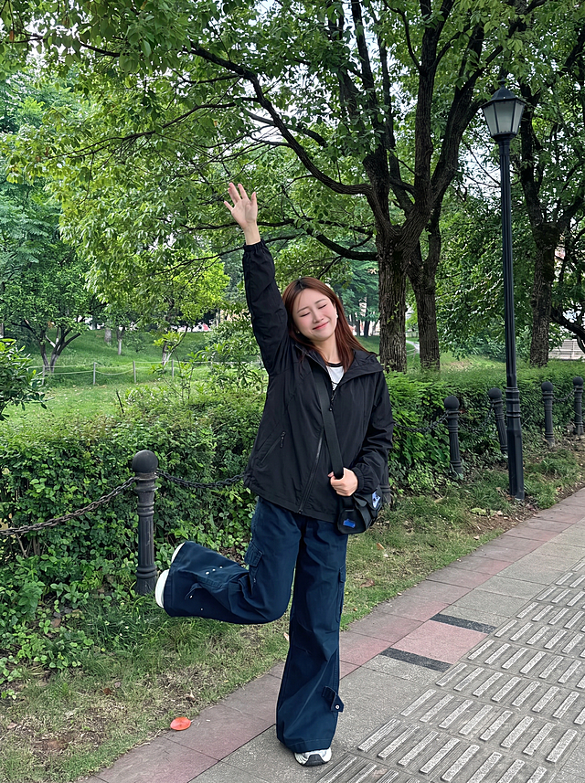 给图1中的女生换上图2中的黑色连帽外套，服装过渡自然平整，保持人物姿势和户外公园背景不变，图片尺寸比例为3比4