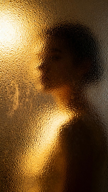 磨砂玻璃质感的半透明画面，金色暖调光源从左侧上方照射，形成明暗对比强烈的光影效果，玻璃表面布满细密凸起的颗粒纹理，光线透过时产生朦胧的散射，画面中隐约可见模糊的人形轮廓，整体呈现出神秘而温暖的氛围，保持磨砂材质的粗糙颗粒感和光线穿透的通透感。预览效果