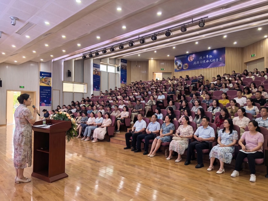 将大会场讲台的讲师换成参考图中穿旗袍的讲师，将阶梯教室所有空座位都坐满，新增入座的人物都穿着夏季服装，包含老师和家长，保持原图整体场景、构图、光线不变预览效果