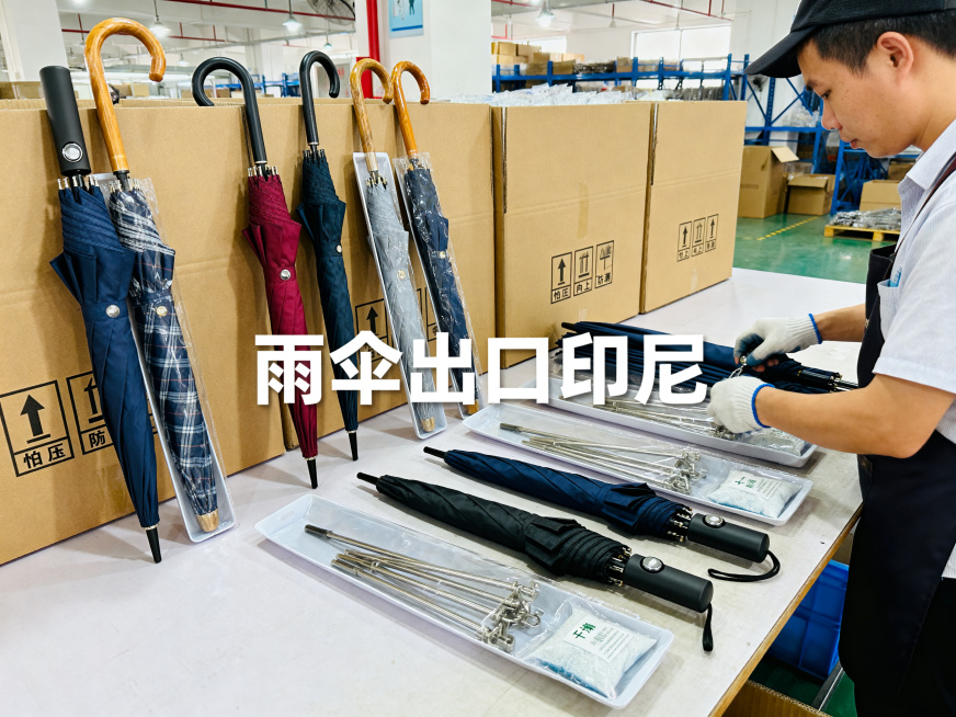 高清晰度商业摄影照片，4:3比例。内容为几款代表性雨伞，包含折叠伞、长柄伞、遮阳伞，操作员正在逐一检查自动伞的弹簧按钮后放入独立包装，骨架含金属件的伞具包装内添加干燥剂，随后整齐顺向码放进贴有“怕压”预览效果