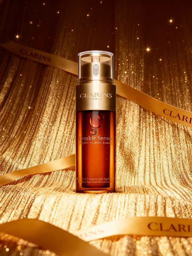 奢华皇室风高端护肤商业摄影风格，华贵璀璨的调性，画面整体采用全金色系（闪耀金、暖金）的高光泽配色，暖调浓郁的奢华光影；采用平视偏轻微仰视视角，主体居中构图；场景为满铺金色闪光线纹肌理的奢华织物背景，搭配印有品牌标识的金色丝带装饰，背景点缀细碎星芒光斑；主体位于画面正中央区域，垂直竖立摆放，占画面约 45% 比例，放置在金色闪光线纹肌理的背景织物之上；主体与环境的关系为：主体置于金色闪光线纹肌理背景，金色丝带环绕呼应，背景的奢华织物肌理与星芒光斑强化华贵层次，暖调高光布光凸显主体的通透金质与精致细节，营造皇室级奢华的护肤产品氛围。