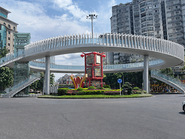 以这张重庆綦江九龙大道环岛街景现状照片为底，保持原图所有元素完全不变：环岛中心花境、红色龙雕塑与"到綦江・如来如愿"标语、路牌、高层住宅楼、行道树、蓝天白云、车流、低机位透视构图均完全保留原样，仅在环岛花境外圈车道外侧上方新增一座单层环形人行天桥。天桥内径与环岛直径相当，圆环正好悬于花境外圈车道外侧上方，体量轻盈真实可建；桥面距地面4.5米、宽约3.5米，桥型为浅灰白色钢结构，桥体上缘为连续的放射状竖向白色格栅，格栅随环体弧度起伏形成柔和包覆膜，内侧为超白玻璃栏板；圆环中部开洞，花境中的红色龙雕塑与城市标语从圆洞正中完整露出；圆环四个象限各伸出一条弧形梯道落至街角，其中一处设通透玻璃垂直电梯井筒；桥墩为简洁圆形钢柱，落于花境外侧绿化带内，车辆照常通行。参考第二张图的天桥格栅与玻璃电梯结构风格。灰绿低饱和色调，建筑细节清晰，8K超高清，真实摄影质感，透视准确，光影与原图一致。