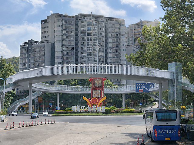 在这张重庆綦江九龙大道环岛街景现状照片基础上，仅新增一座单层环形人行天桥，其余所有现状元素必须完全保持不变：环岛中心花境、红色龙雕塑与"到綦江·如来如愿"标语、路牌、高层住宅楼、行道树、蓝天白云、车流、低机位透视构图全部原样保留。新增环形天桥设计要求：以环岛中心花境为圆心，圆环内径与环岛直径相当，圆环正好悬于花境外圈车道外侧上方，体量轻盈且尽可能大；桥面距地面4.5米（约三个汽车高度），桥宽约3.5米；桥体为浅灰白色钢结构，桥体上缘为连续的放射状竖向白色格栅，格栅随环体弧度起伏形成柔和包覆膜，桥体内侧为超白玻璃栏板；圆环中部开洞，花境中的红色雕塑与城市标语从圆洞正中完整露出；圆环四个象限各伸出一条弧形梯道落至街角，其中一处设置通透玻璃垂直电梯井筒；桥墩为简洁圆形钢柱，落于花境外侧绿化带内，不影响车道，车辆照常通行。整体风格灰绿低饱和色调，建筑体量真实可建，格栅细节清晰锐利，超高清写实摄影质感，8K精细细节，自然光真实光影，透视准确，与原图环境完美融合，看不出修改痕迹。