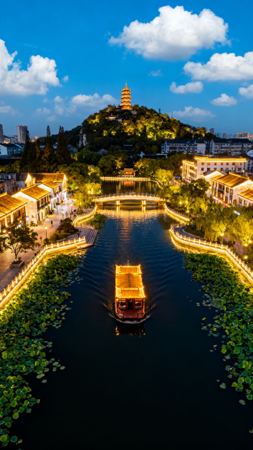 俯视图视角，南通濠河夜景灯火璀璨，游船为视觉中心，远景狼山静谧矗立，蓝天白云春意盎然，河面绿叶悠悠漂浮，画面浪漫动人预览效果