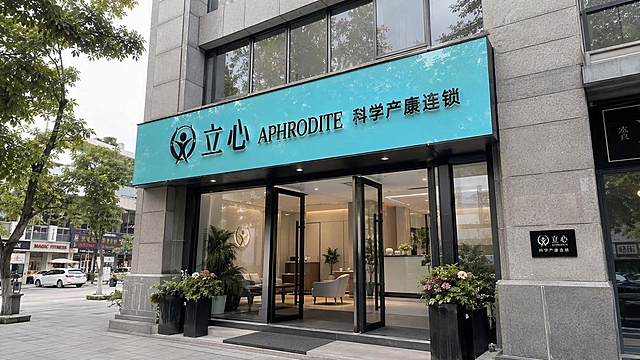 真实街头的高端产康门店门头，非弧形建筑，是方正的现代底商建筑；门头沿用第二张图的样式：青蓝色亮面长招牌、黑色金属立体字“立心 APHRODITE 科学产康连锁”、左侧人形LOGO，LOGO样式完全不变，品牌青蓝色调保持一致，去掉No.006编号；建筑外立面是高级灰色石材，玻璃双开门敞开，门口有精致绿植，墙面有黑色品牌小标识牌，周边是城市高端街区街景，有行道树、整洁的人行道、远处的商铺，白天自然光，轻微仰视角度，真实街拍质感，高清写实，高端大气，生活化不刻意