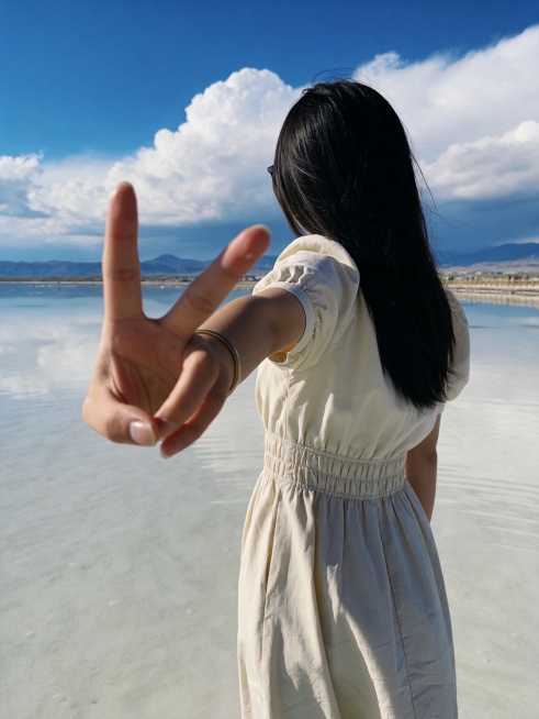 女生游客在茶卡盐湖天空之镜的背影自拍，女生穿浅色连衣裙站在盐湖镜面浅水中，背影对着镜头伸手比耶，远处是蓝色天空和白色云朵，盐湖倒影清晰，氛围感治愈，自然真实游客照风格，竖版适合小红书配图，画质清晰预览效果