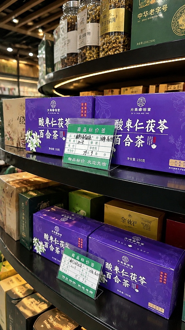 基于上传的原图进行修改，保留原有商品酸枣仁茯苓百合茶包装盒和商品标价签，完整保留标签上的所有文字内容和产品包装上的所有信息，仅更换背景为高端商场的货架，货架上自然摆放和摆满其他商品，营造高级精致的商超零售氛围，整体画面自然真实，光影协调，商品融入场景自然，无抠图痕迹，保持标签内容完全不变