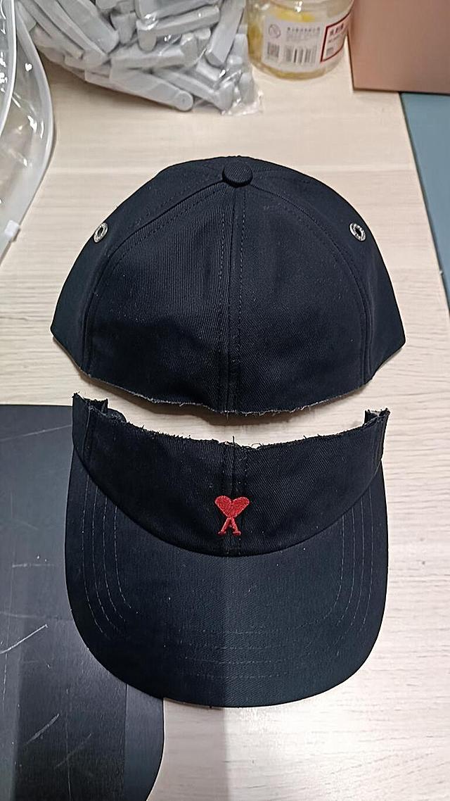 A black six-panel baseball cap with red heart A embroidered logo on a light wood table in a casual indoor background with plastic packages, yellow bottle and boxes, the cap has been cut horizontally with scissors into two separate halves, the cut is a nearly straight horizontal line with natural slight unevenness, NOT zigzag and NOT triangular sawtooth, very minimal loose threads, fresh cut appearance, the upper crown half is separated from the lower cap body half and both pieces are placed near each other, no missing parts, complete fabric is preserved, the hat's original condition and fabric texture preserved, only the hat is modified, the wood table and background objects unchanged, original image composition and size kept, realistic photography, high detail, high definition