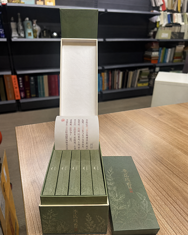 参考图中的包装盒结构和摆放方式完全保留，将包装重新设计为方案七【本草清源】风格。主色调为复古豆沙绿/草药绿色，天然古朴。艾草、甘草等草本植物以精细线条描绘，烫白或暗压。"李氏珍香"字体为极细现代文豪体，烫浅香槟金色。草木纸质感，原生纸浆颗粒感。内部卡片米白底色，左上方"药典"风格红圈印章。大盒内整齐排列5个小盒和1张折叠卡片，旁边放一个独立小盒。保持参考图的拍摄角度、木质桌面、书架背景、自然光效果。产品摄影级真实质感