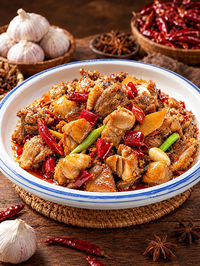 基于参考图中的红烧鸡块菜品，生成侧面微俯角度的美食摄影图，保持菜品色泽红亮、辣椒葱段蒜瓣配料丰富，蓝边白瓷盘装盛，木质桌面背景，背景有独蒜、干辣椒、八角等香料，暖色调专业美食摄影，高清细节，诱人食欲