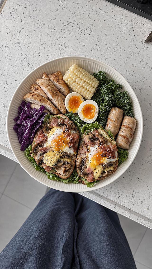 俯视视角，米白色带竖纹的圆盘里装着鸡胸肉蔬菜拼盘，有煎鸡胸肉片、玉米段、羽衣甘蓝、紫甘蓝、水煮蛋、肉卷，还有两片蘑菇培根煎蛋开放式吐司。背景是浅灰色带细斑点台面，画面下方能看到穿着深蓝色裤子的腿。低饱和度ins风滤镜，柔和自然光，暖灰色调，美食摄影，真实质感