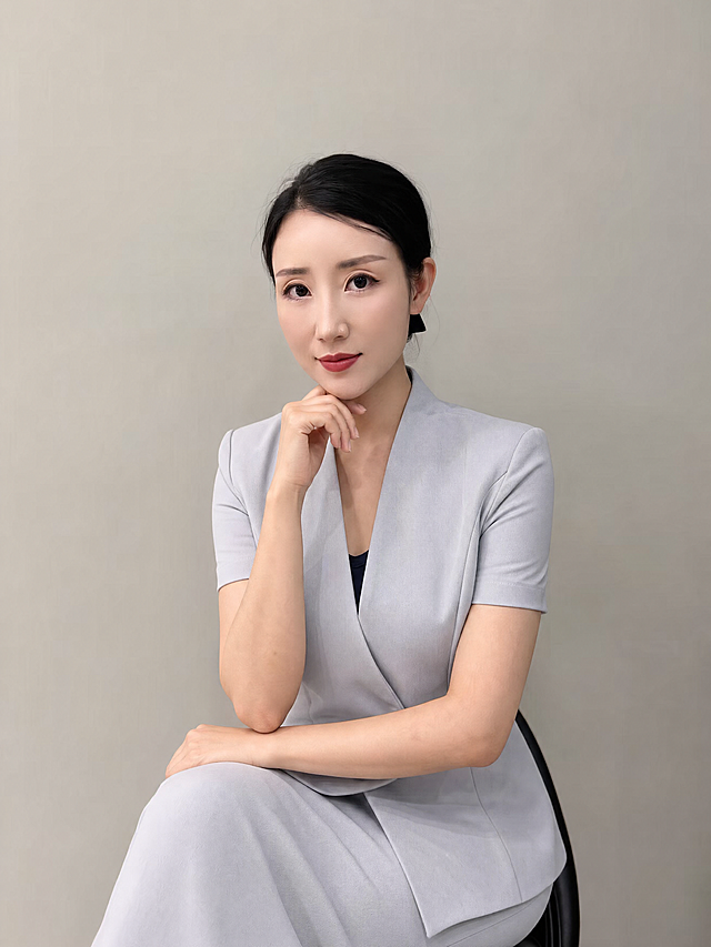 参考图中保留人物脸型、发型、年龄等特征生成职场形象照,标准焦段50mm,f/4光圈(f/2.8-f/4区间),平视视角(镜头与人物视线齐平);人物占画面70%,人物居中(居中构图),位于画面中心,为视觉聚焦点,入境范围头部及以下至臀部下方10cm(半身像);左右留少量背景空间;头部与画面顶部保留5%空间;头部发型完整,头肩比例协调,人物结构准确;
身穿浅灰色高支精梳棉职业西装外套，(v领设计,面料挺括细腻);内搭深色打底衫;下装同材质浅灰色西装套裙;着装正式干练;亲和专业;身形匀称,姿态端正,身体挺拔。呈自然坐姿，手扶下巴，肢体放松且自信;
发质微微柔顺，微微蓬松;
简约微微暖调暖灰米杏纯色背景(呈现均匀柔和质感),背景光线均匀柔和,强化色彩对比的高级感,极简干净;
画质超高分辨率、细节清晰,现代精致感风格;肤质精细化处理;眼神直视镜头,目光坚定且澄澈,表情自然。嘴角上扬;
清透自然裸妆(轻薄底妆+淡唇妆)妆感轻薄轻透裸肌感,通透感肤质,肤质自然细腻,肤色健康均匀,雾面柔光裸肌感,保留真实皮肤细节｡正面柔光补光,4800K中性光,面部光比1:1.5,色调明亮｡中近景写实摄影,对焦清晰,高清保留皮肤细节｡
分辨率4K,去除人物之外的原配饰，保留干净画面。严格按照图片内容生成。
画面比例3:4
