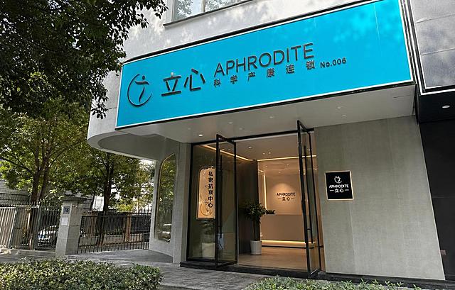 参考图中的门头品牌LOGO、品牌名称文字“立心 APHRODITE 科学产康连锁”、品牌青蓝色调完全保持不变，更换为完全不同的真实门头外观，不同的建筑外立面结构、不同的门头造型、不同的周边街景环境、不同的拍摄角度和光线，真实街景实拍质感，高清写实，看起来是不同的门店，不像同一个门头