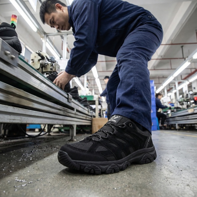 工厂流水线场景，穿着深蓝色工作服的男性工人正在工作，脚上穿着这双黑色户外徒步鞋，采用低视角拍摄，画面重点突出脚上的鞋子，鞋子细节清晰，车间光线明亮预览效果