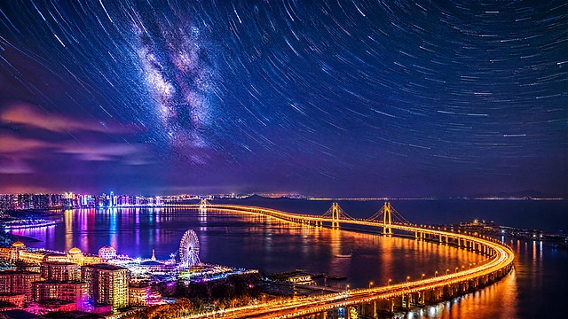 画面上半部分是深邃的星空延时，繁星点点，银河清晰可见，星轨流动感；下半部分是星海湾跨海大桥的夜景灯光，大桥蜿蜒横跨海面，桥面和桥塔点亮暖金色灯光，海面倒映着霓虹色彩。整体配色为蓝紫色调为主，搭配暖金色大桥灯光和霓虹灯光色彩，营造嘉年华般的热闹氛围，微醺体验的浪漫感，视听盛宴的震撼视觉效果，展现滨海夜生活的繁华与活力。电影级质感，高细节，夜景摄影风格，光影层次丰富。