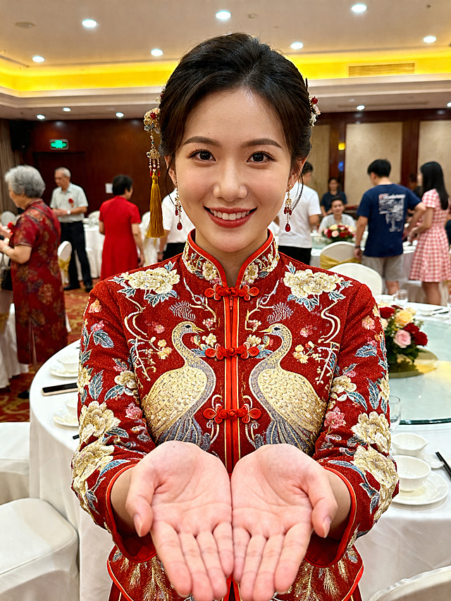 画面中心是一位年轻东亚女性，身穿精致红色中式秀禾服，衣服绣有金色凤凰、花卉与传统纹样，盘扣细节清晰；人物自然微笑，表情温婉亲切，不夸张咧嘴，双手在胸前呈标准持红包姿势，双手掌心相对、呈捧物状，双手之间预留出平整的矩形空白区域用于放置红包，手部结构自然比例正常无畸形；妆容清透自然，佩戴中式流苏耳饰与发簪，发型精致。场景是真实中式婚宴现场，酒店宴会厅内景，暖黄色柔和室内灯光；背景可见铺白色桌布的圆桌、摆放整齐的餐具、桌花，宾客穿着日常喜庆服饰，长辈穿中式礼服、年轻宾客穿休闲正装和连衣裙，背景人物自然活动不突兀，整体氛围喜庆温馨，符合真实婚礼场景。风格参考真实手机原相机拍摄质感，无过度磨皮，无AI感，保留皮肤自然纹理、毛孔细节，色彩还原真实，光影自然柔和，带生活纪实感，高清8K，细节清晰，无过度美颜，像真实婚礼跟拍的手机直出照片。构图是中近景平视构图，人物居中，聚焦人物上半身与手部，背景轻微虚化突出主体，画面层次分明，符合日常拍照的视角，无夸张透视。