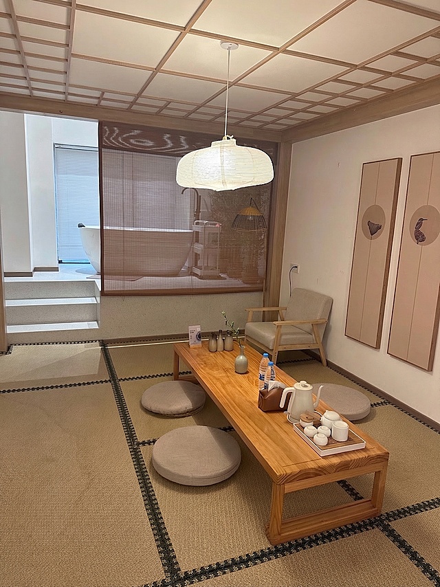 在墙角空地上添加一个小型单人沙发，在原木长桌空闲位置放置一个简约小花瓶，保留原有的整套白瓷茶具，保持原有日式禅风茶室风格不变，整体协调自然