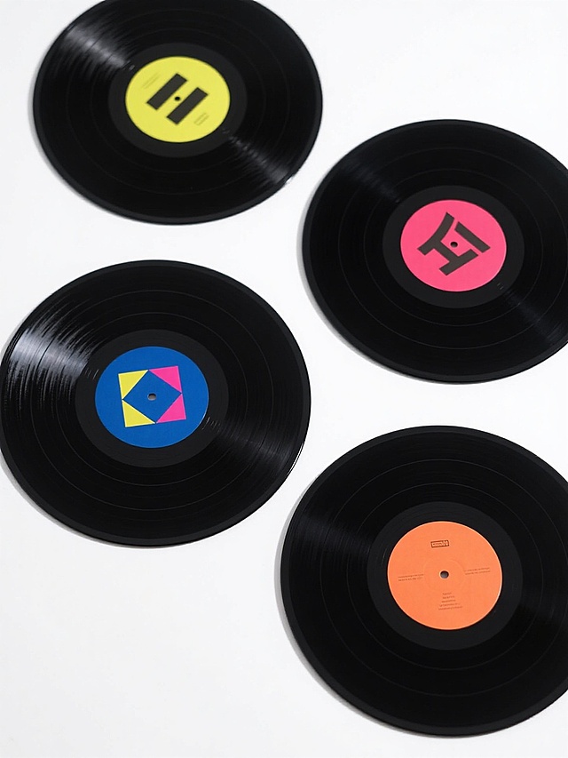 白色背景上排列整齐的四张不同样式不同配色的黑胶唱片，每张唱片都有精致的黑色同心圆纹路和光滑反光的表面质感，中心标签分别采用不同鲜艳颜色并带有简约几何图案设计，整体呈现出干净利落的现代产品摄影风格，光线均匀柔和，阴影细腻自然，构图平衡对称，突出唱片的材质细节和色彩对比。