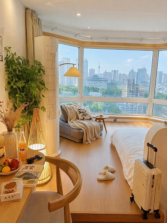 基于原图西安民宿弧形落地窗城景房场景进行优化重绘：整体色调明亮通透，不压暗，避免阴沉发灰；滤镜不过黄，保留原图亮度和自然暖木色调，画面真实协调，窗外西安城市景观清晰明亮。去掉所有人物，只保留纯净场景。增强西安民宿生活感和入住感，丰富摆件物品：在房间角落或床边放置一个浅色（米白色）行李箱，懒人沙发上搭一条针织毛毯和一个抱枕，旁边小桌上放一杯茶、一本西安旅游攻略、一副墨镜，绿植旁加一个小花瓶插着干花，书桌上增加西安特色糕点、冰峰饮料、水果盘，地上放一双拖鞋，体现西安古城民宿真实入住的氛围，物品摆放自然不繁杂，画面和谐有氛围感。小红书民宿风格，真实感强，4K高清，独立出图不拼图，3:4竖版比例。