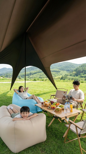 一家三口在茂盛的绿草坪上，在一个大型咖啡色内部黑色遮阳棚下享受夏季露营旅行，视角在帐篷内部背景是起伏的绿色山丘。一位年轻女子躺在浅蓝色充气沙发上，双手放在头后。一个小男孩躺在米白充气沙发上，朝镜头看。预览效果