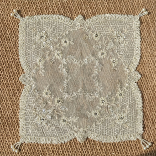 严格还原30×30cm菱形蕾丝餐垫产品细节，米白色网纱基底，带有金线花卉刺绣，水钻花蕊，四角珍珠流苏坠饰，餐垫在浅棕色编织地毯上平铺展开，俯视平铺构图，餐垫完整展示无遮挡，画面风格简洁大气、轻奢优雅，预览效果