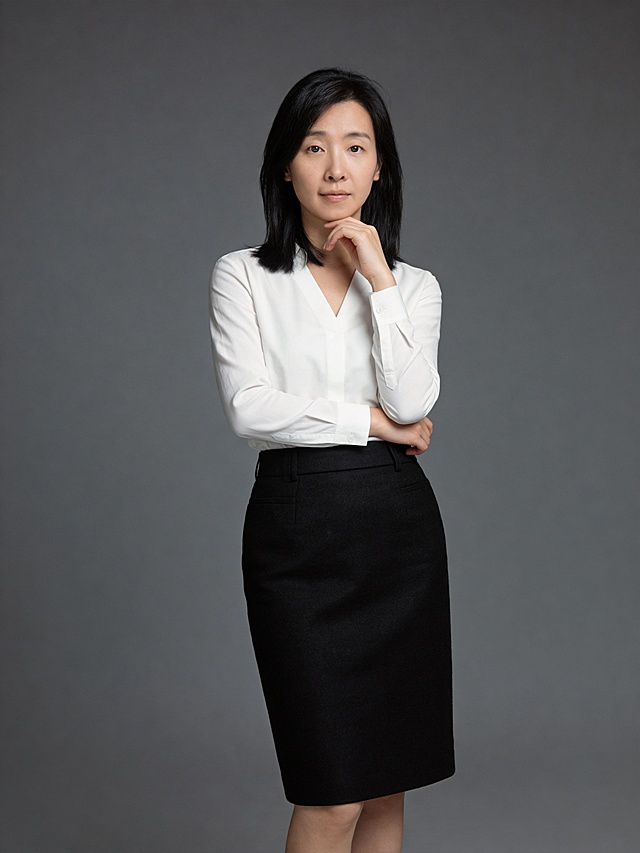 参考图中保留人物脸型、发型、年龄等特征生成职业形象照，身着净白色V领韩式长袖衬衫，优选细腻棉质面料，自带柔和垂坠感，V领弧度修饰颈部线条，袖口微收利落不拖沓；下搭高腰黑色哑面羊毛混纺材质包裙，高腰线设计，通勤感。标准焦段50mm，F/4光圈，处于F/2.8-F/4区间，平视视角，镜头与人物视线齐平；人物占画面70%，人物居中，居中构图，位于画面中心，为视觉聚焦点，入境范围头部及以下至臀部下方10cm，半身像构图。左手轻托下颌，右手自然交叉于臂弯，身姿随性有度，自然松弛姿势。简约冷调深灰色纯色背景，呈现均匀柔和质感，极简干净背景，背景光线均匀柔和，强化色彩对比的高级感。画质超高分辨率，细节清晰，现代精致感风格；肤质精细化处理，眼神直视镜头，目光坚定澄澈，表情自然。肤色均匀，柔光美白细腻陶瓷肌肤，哑光肤质，肤色均匀提亮；杰作，最佳画质，超高细节，写实摄影，中近景人像，皮肤细腻通透，白皙清透皮肤，柔光，正面补光，画面干净清新，高清皮肤细节，面部光比1:1.5，4800K中性光，微微修饰脸型，肤质哑光细腻，面部雾面柔光，轻透裸肌感，真实毛孔纹理，分辨率4K