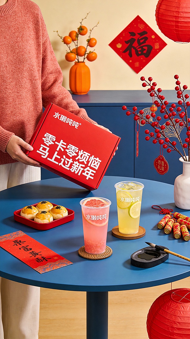 由穿米红色毛衣的人物手持倾斜展示；背景搭配新年装饰（红果枝、橘色挂饰、红色福字）与红色托盘装着蛋黄酥，置于蓝色圆桌，桌上还有2杯透明杯装果茶饮品（粉色西柚汁、柠檬汁）2杯饮品比例大小保持一致，桌上还有对联，毛笔、砚台点缀画面；构图采用生活场景式布局，礼盒为视觉核心，装饰元素烘托新年氛围，人物肢体增强互动感；视觉效果呈现新年温馨礼品风格，整体呈现为新年健康礼品推广场景，强化温馨热闹的节日氛围与产品的生活感，传递出新年赠礼的温暖调性。春节喜庆氛围