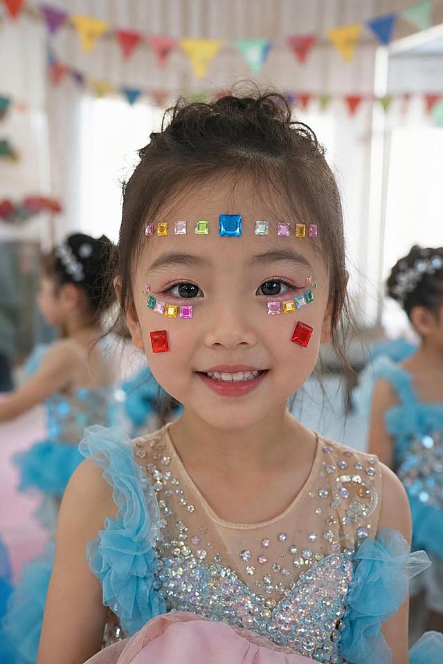 2:3竖版真实手机生活照，正面略俯角度，幼儿园小舞台。小女孩正对镜头微笑，脸上方形钻贴夸张几何感：眉心一颗大蓝色方形钻，两眼周围各贴一排大小方形钻像几何眼影，双颊各贴一颗大红色方形钻，额头横向也贴几颗方形钻连成线，整体闪亮利落、像几何舞台妆。方形钻的形状、刻面、通透质感、颜色严格还原参考图中的方形钻贴产品。背景虚化教室布置、彩色三角旗、其他小朋友，午后自然光、像老师随手拍、真实手机质感、非影棚，无文字水印。小女孩穿演出服，淡妆，皮肤自然，画面真实感强。