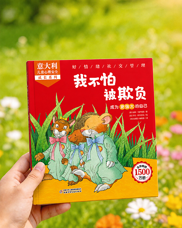 一只手拿着图2中的书籍《我不怕被欺负》，将原本图1的书籍替换成图2的书籍，保持背景绿色草地鲜花不变，保持构图不变