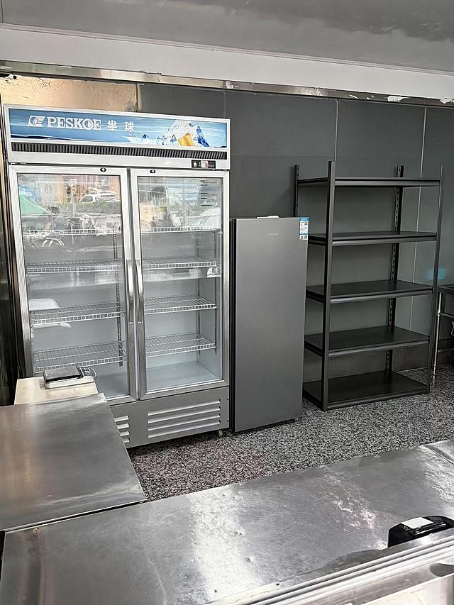 去除右侧5层金属货架上的所有物品（包括篮子、锅、塑料盒、纸箱、包装袋等所有摆放在货架上的物品），仅保留空的货架主体结构，货架的金属框架和层板完整保留。同时将左侧双门透明玻璃展示冰箱内部的所有物品全部清空，仅保留冰箱内部的金属层架和空的内部空间，冰箱玻璃门、外观结构完全保留。严格保持房间内其他所有主体、细节、结构、颜色、光线、透视角度完全不变，不得添加或修改任何其他元素。去除物品后的区域用周围真实的墙面、地面、货架结构和冰箱内壁自然填充，光影和材质与原图一致，过渡自然无痕。最后整体提升图片清晰度，画质自然真实，保持原照片的真实感。处理效果与同场景另一角度照片保持一致。
